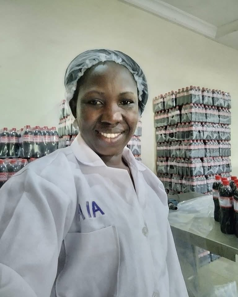 A smiling female factory worker in a white lab coat and hairnet standing in front of stacked soda bottles.